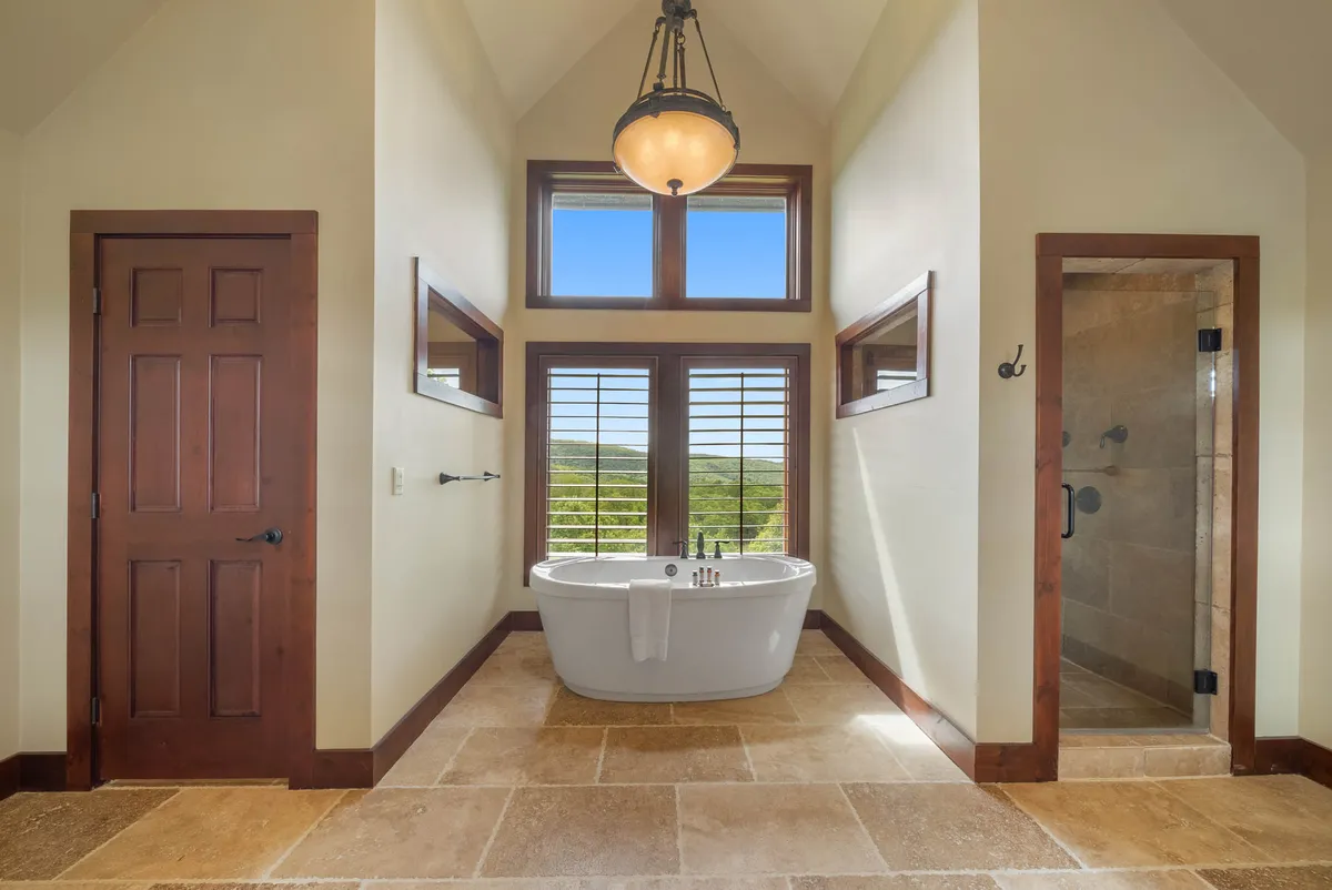 A spacious bathroom with a freestanding soaking tub positioned in front of large windows offering a scenic view. The area includes a separate shower and wooden door, exuding tranquility and luxury—ideal for indulgent relaxation after a long day.