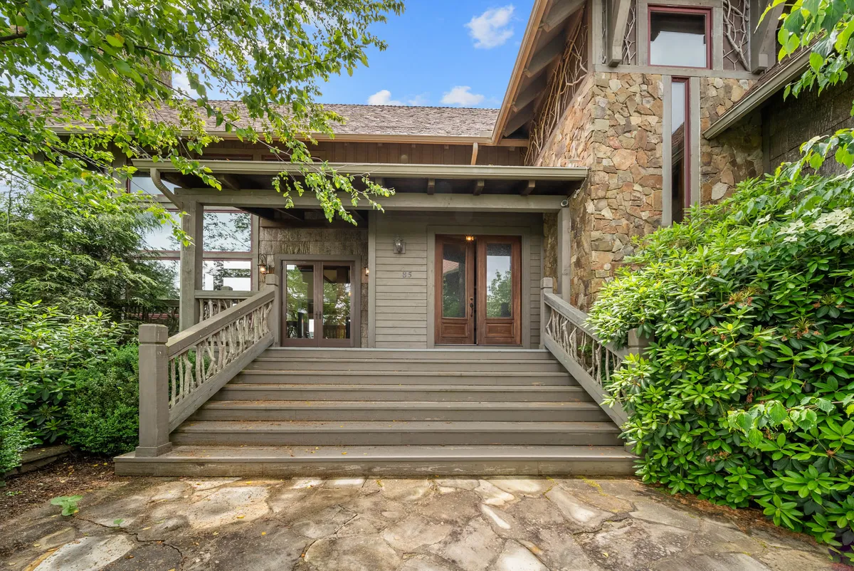 This photo features wooden steps leading to double doors, a stone facade, and vibrant green landscaping. The scene exudes warmth and invites visitors to gather, relax, and enjoy the natural surroundings.