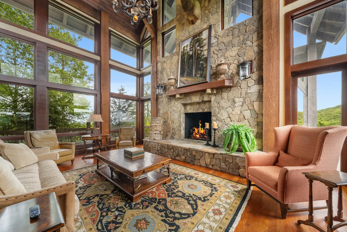 A spacious living area anchored by a large stone fireplace, elegant chandelier, richly patterned rug, and expansive windows revealing a beautiful exterior view. The room's arrangement encourages relaxation and family gatherings.