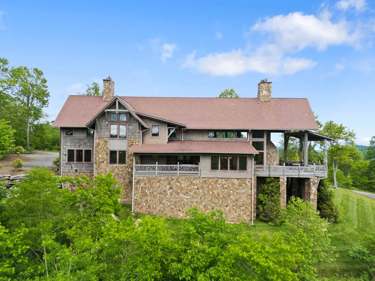 A tranquil exterior shot highlighting the home's stone façade, rustic wooden beams, large windows, and lush greenery. The porch invites outdoor relaxation and gatherings, blending the comfort of home with the beauty of surrounding nature.