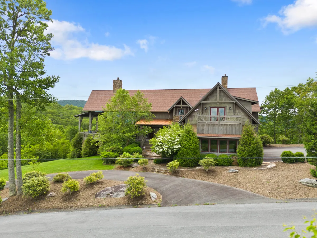 A welcoming exterior shot of the home nestled among mature trees, with a well-maintained driveway, landscaped garden, and the porch visible. The surrounding natural setting hints at immediate access to trails and outdoor spaces, while the elevation offers a sense of seclusion and tranquility.
