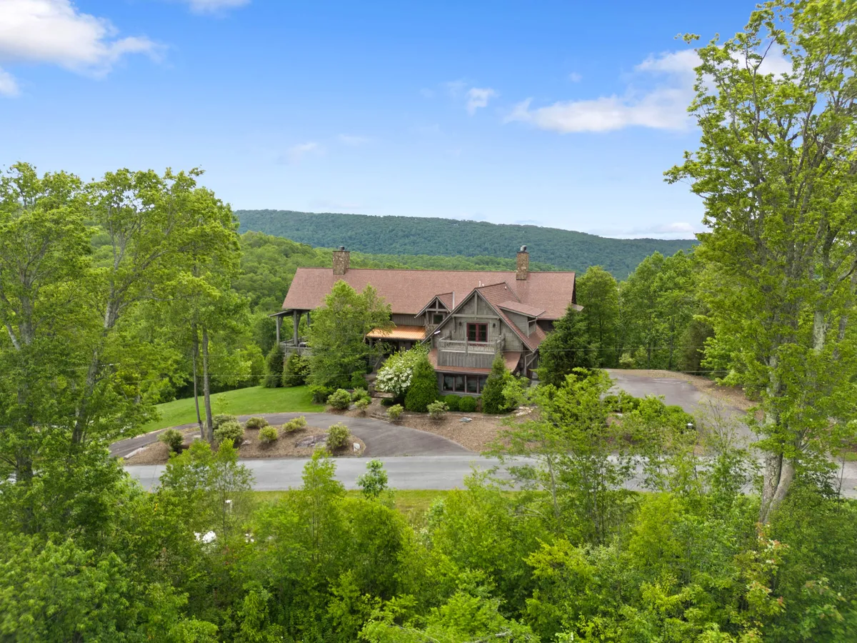The image showcases a stunning, spacious luxury mountain home nestled among lush trees, with a sweeping driveway leading up to its grand entrance. The sun is setting over rolling hills in the background, casting a warm golden glow, hinting at breathtaking sunset and stargazing opportunities from the property.