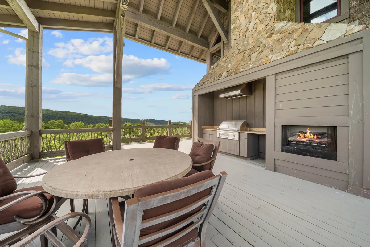 An inviting exterior scene presenting a circular wooden dining table and comfortable seating on a covered terrace, accompanied by an outdoor kitchen with a built-in grill and a prominent stone fireplace. The space is oriented for sunset viewing and group celebrations, with natural surroundings visible beyond the deck.