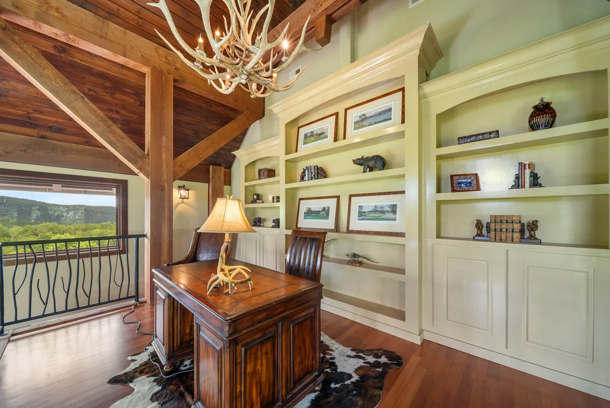 A cozy loft office space featuring a wooden desk positioned by a large window with scenic views, an antler chandelier overhead, and built-in shelves decorated with books and ornaments. Natural light fills the workspace, creating an inspiring environment.