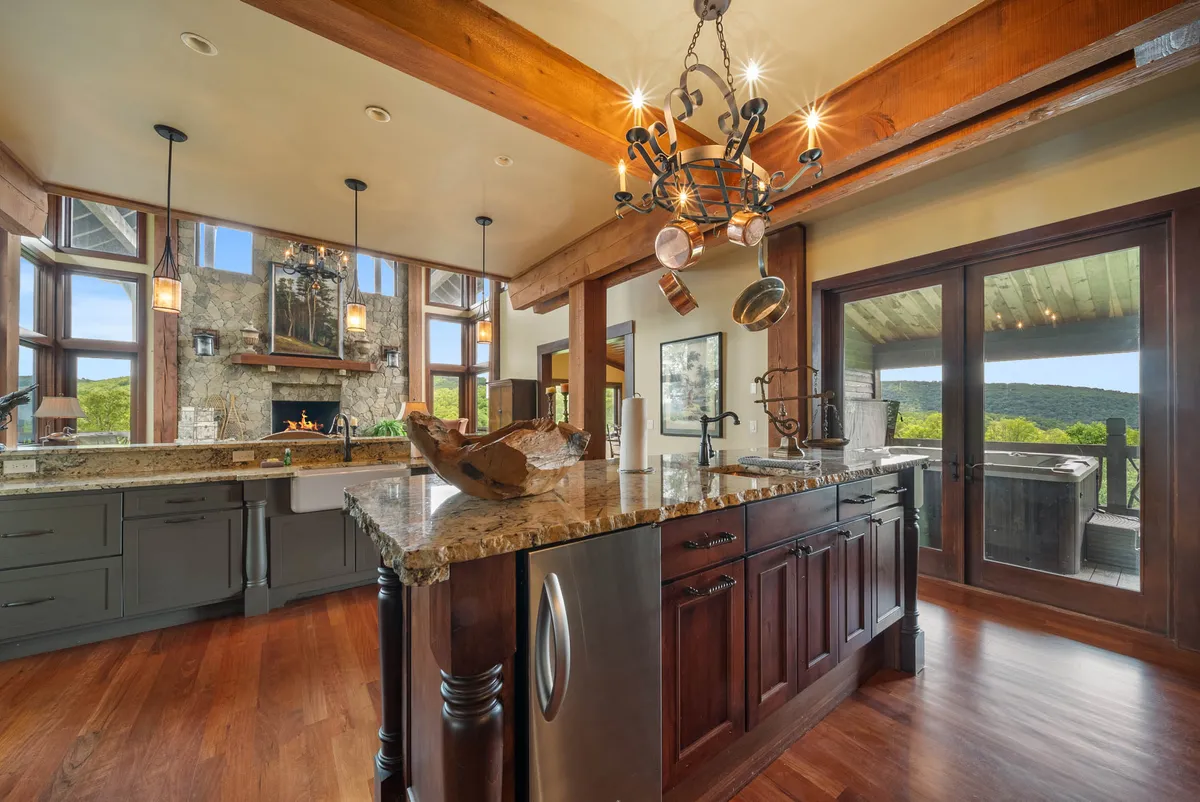 A beautiful kitchen scene featuring a granite countertop, exposed wooden beams overhead, large windows letting in natural light, and an elegant chandelier. The space is clearly designed for culinary experiences and socializing, with an open-plan that visually connects to adjoining living areas.