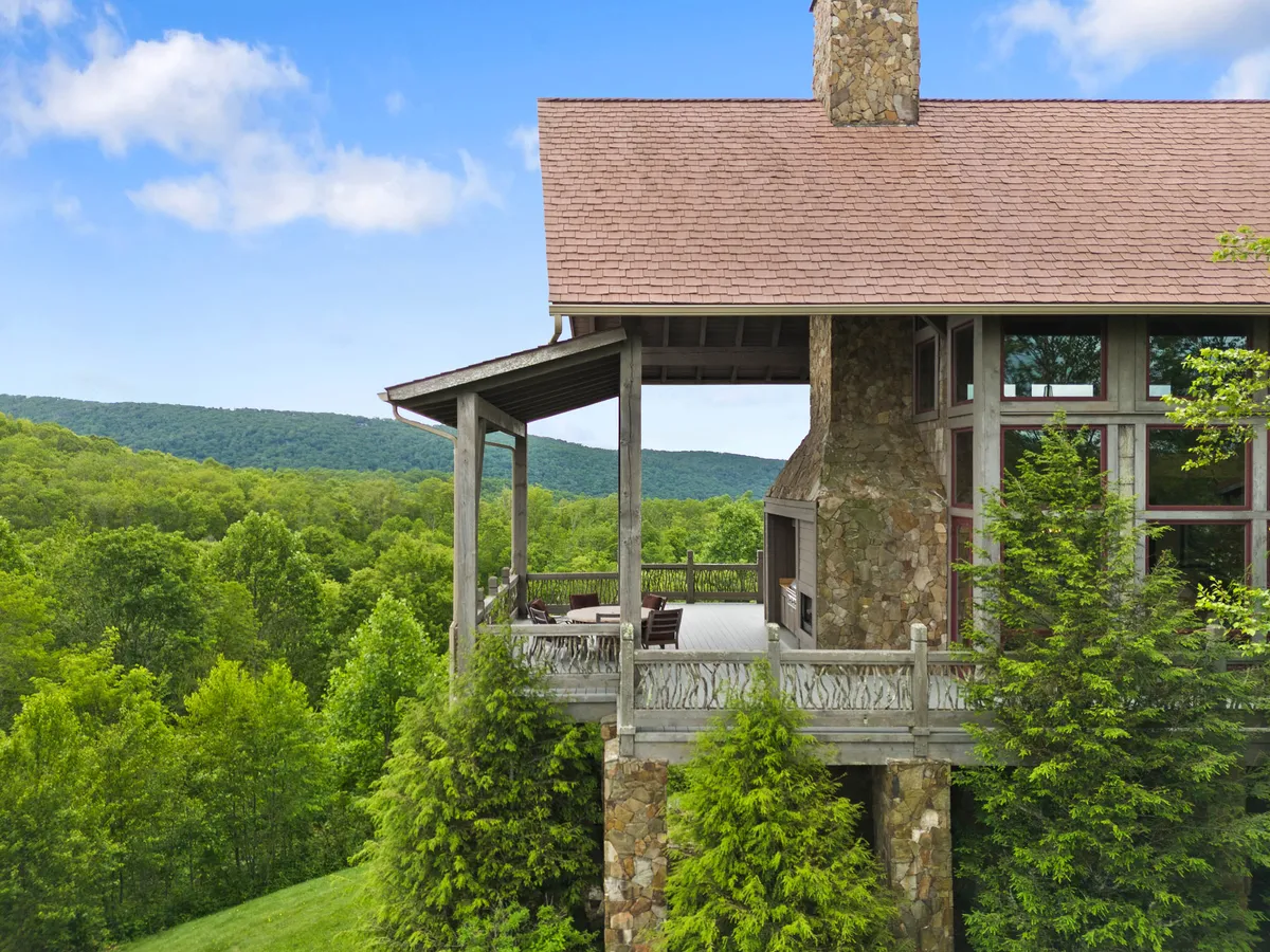This exterior image captures the home's impressive stone exterior and wooden deck, surrounded by lush greenery with expansive mountain views stretching into the distance. The composition highlights the seamless blend of luxury living with the natural landscape, inviting guests to relax on the deck and take in the panoramic scenery.