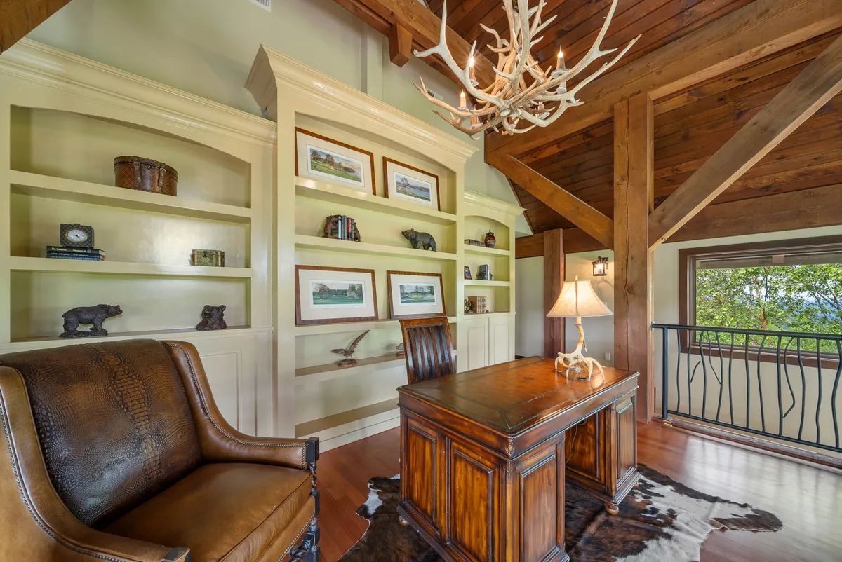 An inviting loft workspace with a wooden desk, leather armchair, antler chandelier, and bookshelves tastefully decorated. A large window provides ample light and a view of the outdoors, emphasizing the room's warmth and inspirational quality.