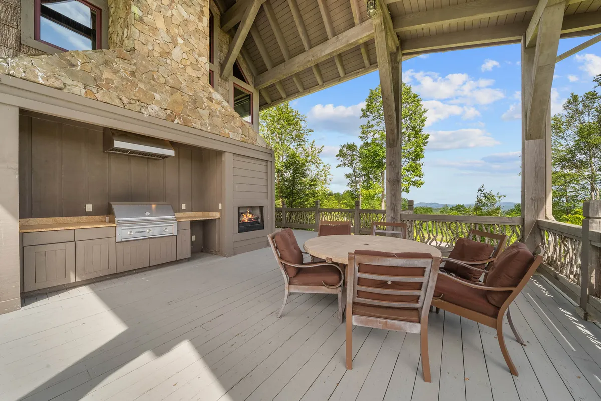 This image showcases a spacious and inviting outdoor living area featuring a built-in grill, a large dining table, a striking stone fireplace, and sturdy wooden beams overhead. The setup is clearly designed for relaxation, entertaining, and enjoying panoramic views, with the partially covered terrace creating a cozy yet open-air atmosphere.