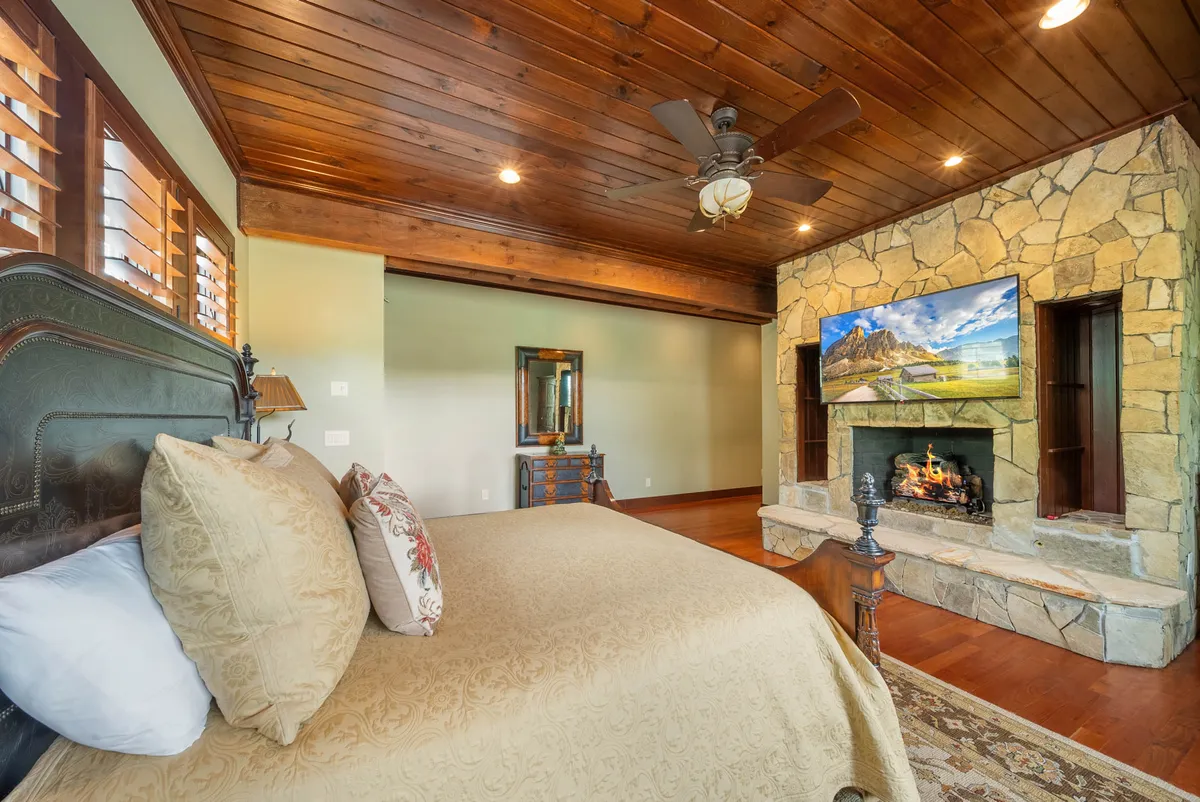 A warm and tranquil king bedroom featuring an ornate bed with decorative pillows, a stone fireplace with a TV above, and classic wooden furniture. The ambiance is cozy and private, with natural light enhancing the inviting mood—ideal for both restful sleep and leisurely evenings.