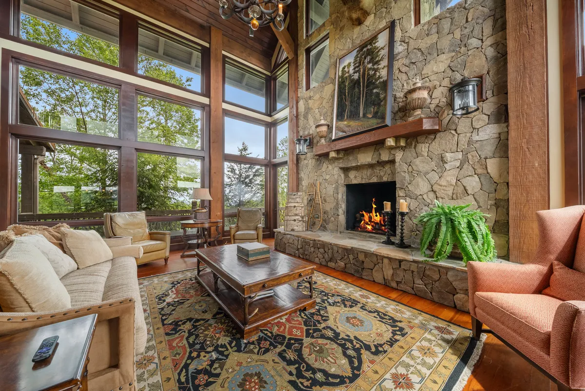 This image showcases a grand interior with a prominent stone fireplace, large windows that reveal sweeping mountain views, exposed wooden beams, an oriental rug, and plush, comfortable seating arranged for gatherings. The ambiance is warm and welcoming, blending rustic elements with luxurious comfort.